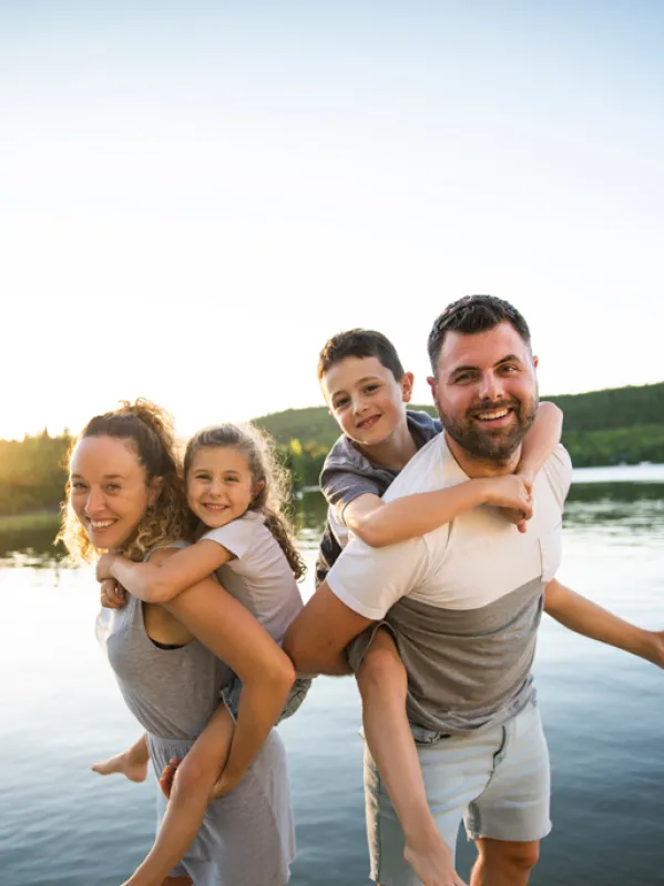 Eine vierköpfige Familie steht in einem See bei Sonnenuntergang und lächelt in die Kamera, die Eltern tragen die Kinder Huckepack.