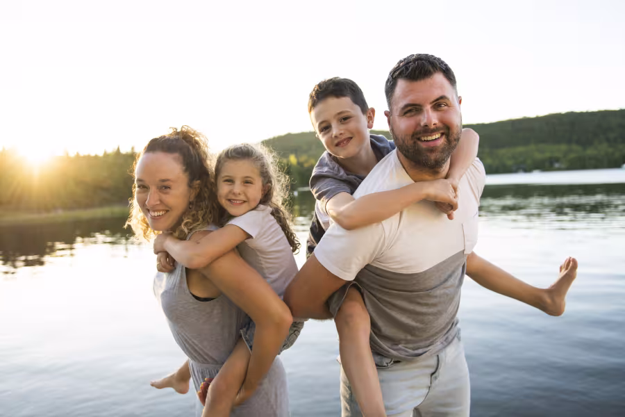 Eine vierköpfige Familie steht in einem See bei Sonnenuntergang und lächelt in die Kamera, die Eltern tragen die Kinder Huckepack.