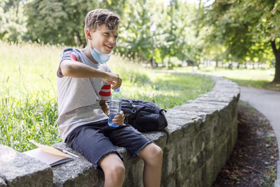 Ein Junge sitzt auf einer Mauer im Schatten und öffnet eine Wasserflasche.